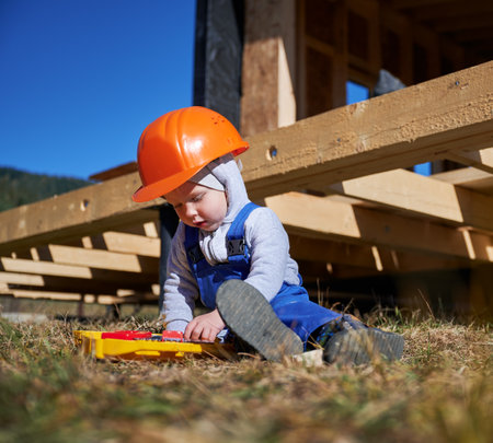Boy toddler playing as builder on construction site. Child carpenter in orange helmet and blue overalls learning to build wooden frame house outdoor on sunny day. Carpentry and workshop concept.の写真素材
