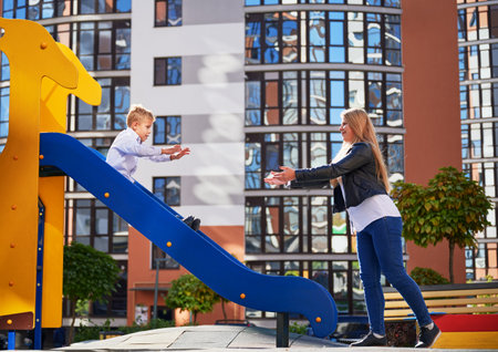 Pretty mum catching male kid, riding on slide at modern courtyard of city residential high-rise buildings. Mother catching little son while standing near slide on playground. Concept of parenthood.の写真素材