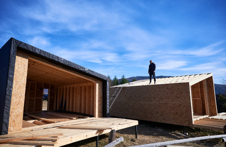 Inspector building wooden frame house on pile foundation in Scandinavian style barnhouse. Bald man standing on construction site, inspecting quality of work on sunny day with blue sky on background.の写真素材
