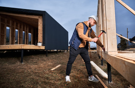 Man worker building wooden frame house on pile foundation. Carpenter hammering nail into wooden truss, using hammer. Carpentry concept.の写真素材