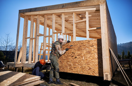 Carpenters hammering nail into OSB panel on the wall of future cottage. Men workers building wooden...