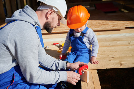 Father with toddler son building wooden frame house. Male builder learing kid to use clamp on construction site, wearing helmet and blue overalls on sunny day. Carpentry and family concept.の写真素材