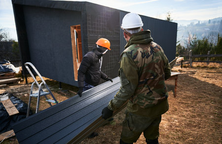 Male builders installing black corrugated iron sheet used as facade of future cottage. Men workers...