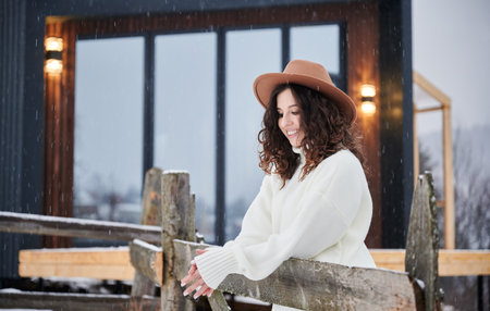 Curly young woman resting near modern barn house in the mountains. Happy female tourist enjoying winter holidays in new cottage.の写真素材