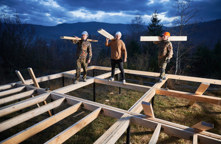 Men workers building wooden frame house on pile foundation. Portrait of three carpenters carrying wooden planks for timber framing. Carpentry concept.の写真素材