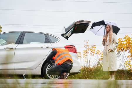 Beautiful woman holding umbrella and looking at roadside assistance worker while man unscrewing lug nuts on car wheel. Auto mechanic repairing woman car on the road. Concept of emergency road service.の写真素材