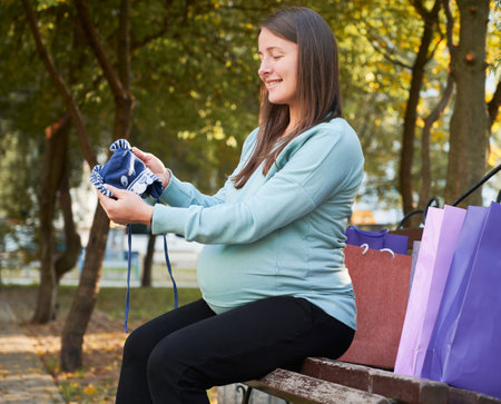 Cheerful pregnant woman with wear for infant feeling carefree. Happy mother with paper bags resting...