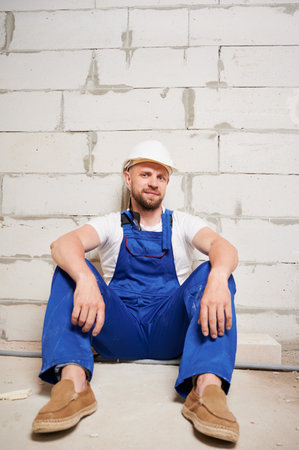 Male builder in work overalls sitting on the floor in building under construction or renovation. Bearded man in safety helmet leaning against brick wall and looking at camera.の写真素材
