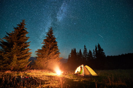 Night camping in mountains under starry sky and Milky way. Female tourist sitting in tent entrance in campsite, admiring landscape and burning campfire. Concept of traveling and hiking.の写真素材
