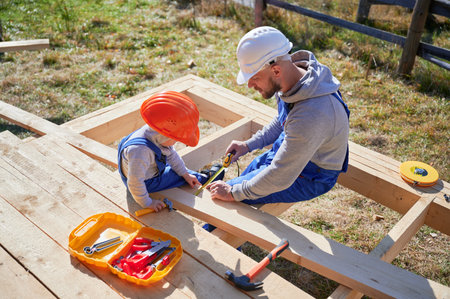 Father with toddler son building wooden frame house. Boy helping his daddy, playing with tape measure on construction site, wearing helmet and blue overalls on sunny day. Carpentry and family concept.の写真素材