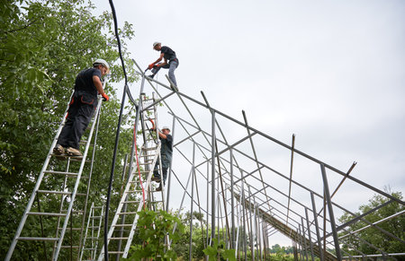 Three men in workwear installing metal poles and rails for photovoltaic solar panels. Male workers using ladders and drill while mounting support structures for solar panels.の写真素材