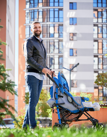 Loving father spending time with newborn kid outside. Side view of young bearded man in casual wear walking with baby stroller on street, with high-rise buildings on background. Concept of parenting.の写真素材