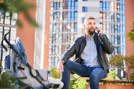 Father making business call, while spending time with baby outdoors. Bearded man holding mobile phone, calling, while baby sleeping in pram at modern courtyard of city residential high-rise buildings.の写真素材