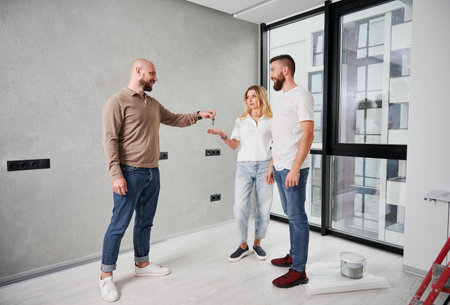 Full length of male realtor handing keys from new apartment to man and woman. Couple standing by the window and receiving keys to new home from real estate agent.の写真素材