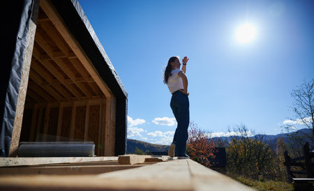 Happy woman on construction site standing on terrace at unfinished wooden frame house on sunny day.の写真素材