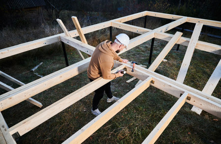 Man worker building wooden frame house on pile foundation. Carpenter installing wooden board, using hammer and screwdriver. Carpentry concept.の写真素材