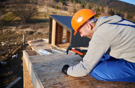 Carpenter hammering nail into OSB panel on the roof top of future cottage. Man worker building wooden frame house. Carpentry and construction concept.の写真素材