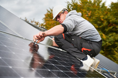 Man builder mounting photovoltaic solar panels on roof of house. Engineer in helmet installing solar module system with help of hex key. Concept of alternative, renewable energy.の写真素材