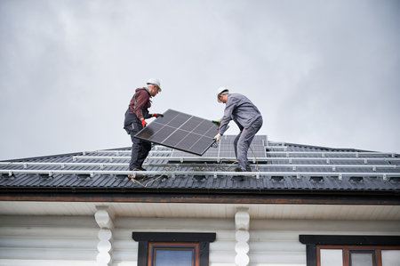 Mounters building solar panel system on roof of house. Men workers in helmets carrying photovoltaic solar module outdoors. Concept of alternative and renewable energy.の写真素材