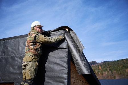Male builder installing waterproof membrane on the wall of future cottage. Man worker building wooden frame house with blue sky on background. Carpentry and construction concept.の写真素材