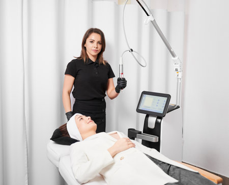 Female cosmetologist holding radiofrequency device and looking at camera while standing by client in cosmetology clinic. Woman receiving rejuvenation skincare treatment in beauty salon.の写真素材