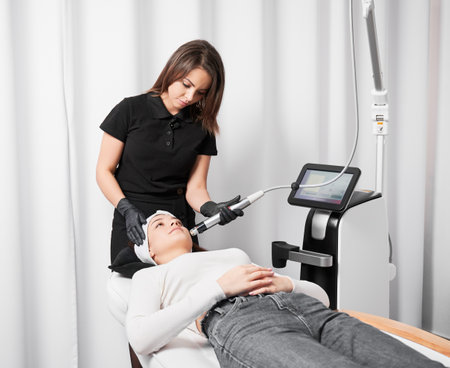 Female cosmetologist using radiofrequency microneedling device while performing face lifting procedure for patient. Woman lying near cosmetology equipment and receiving skincare treatment.の写真素材
