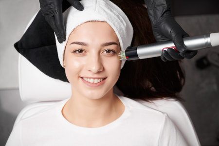 Top view of medical esthetician using radiofrequency device while performing facial treatment in cosmetology clinic. Smiling woman wearing headband while having rejuvenation skincare procedure.の写真素材