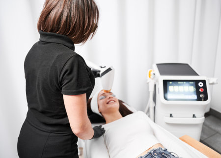 Cosmetologist using diode laser device while performing rejuvenation facial treatment. Woman lying near cosmetology equipment while having face lifting procedure. Focus on female doctor.の写真素材