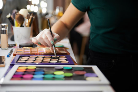 Close up of female hand in sterile gloves holding cosmetic makeup brush above eyeshadow palette. Makeup artist standing by the table with professional cosmetics in beauty salon.の写真素材