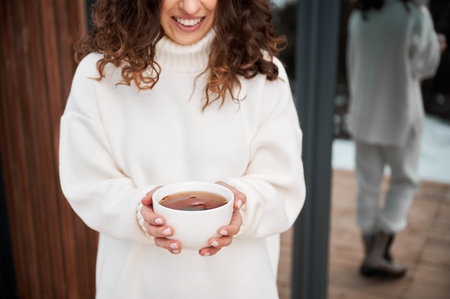 Close up of happy female tourist holding cup of tea, enjoying in new cottage in winter. Curly young woman resting on terrace of modern barn house in the mountains.の写真素材
