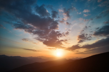 Panoramic view of bright evening sun and dramatic cloudy sky over mountains. Magnificent scenery of mountain hills under colorful evening sky with clouds and setting sun. Beauty of nature concept.の写真素材