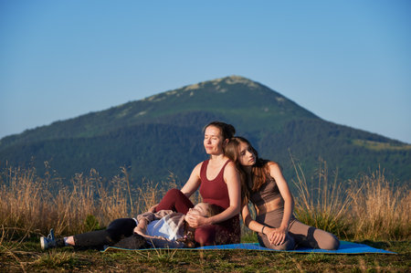 Young mother meeting sunrise with her daughters. Relaxing after morning workout. Three girls sitting on mat on background of mountains.の写真素材