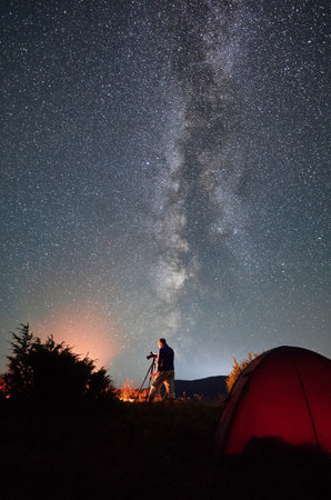 Professional photo shoot of Milky Way in mountains. Male photographer taking photos of starry night in mountains. Back view of man with camera between next to his tent.の写真素材