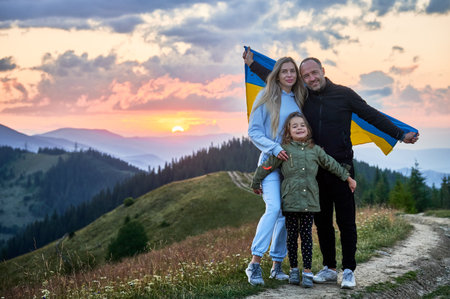 Happy family of hikers waving Ukrainian flag. Man with his wife and daughter standing with symbol of invincibility on background of Carpathian mountains at sunset.の写真素材