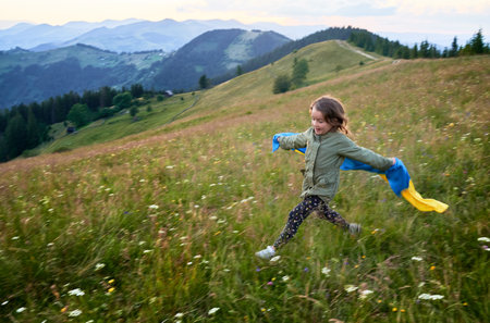 Cute girl running in mountains with Ukrainian flag. Little girl smiling at camera with Ukrainian flag on her shoulders. Feeling freedom in young people.の写真素材