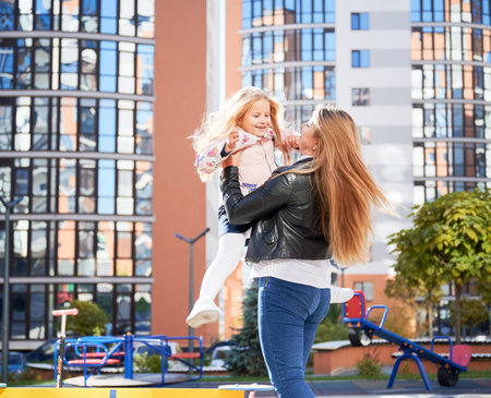 Pretty female hugging adorable daughter, spinning around with blowing hair at playground. Side view of cute little girl having fun with attractive blond mother outdoor. Concept of childhood.の写真素材
