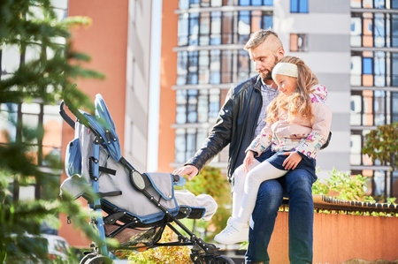 Loving father spending time with newborn kid outside. Side view of young bearded man in casual wear walking with baby stroller on street, with high-rise buildings on background. Concept of parenting.の写真素材