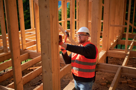 Carpenter constructing wooden frame house. Bearded man with glasses is hammering nails into the structure, wearing protective helmet and construction vest. Concept of modern ecological construction.の写真素材