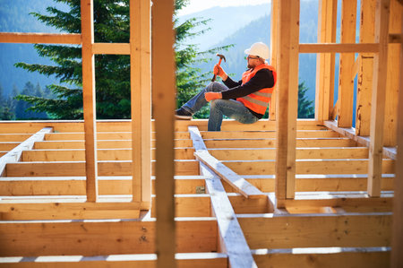Carpenter constructing wooden frame house near the forest. Bearded man in glasses holding hammer, dressed in protective helmet and orange safety vest. Concept of ecological modern construction.の写真素材
