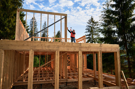 Wooden two-story, frame-built home is being erected by the carpenter near the forest. Man is driving in nails with hammer. Underpinning idea of this venture is modern, ecological building practices.の写真素材