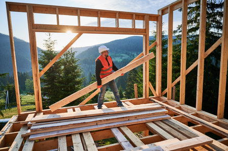 Carpenter constructing wooden-framed house near the woods at sunset. Man holding large joist and dressed in work clothes and helmet. Concept of modern and environmentally-friendly construction.の写真素材
