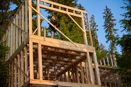 Carpenter constructing wooden framed two-story house near the forest. Bearded man with glasses holds hammer, dressed in protective helmet. Concept of environmentally friendly modern construction.の写真素材