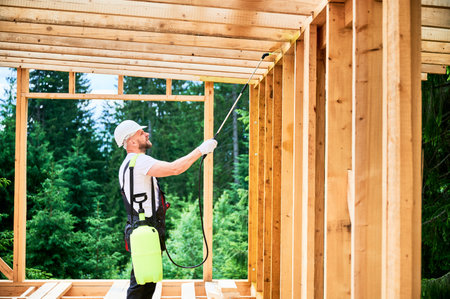 Laborer constructing wooden frame house near forest. Man treating woods, applying fire retardant using sprayer, while dressed in protective suit, helmet. Concept of modern eco-friendly construction.の写真素材