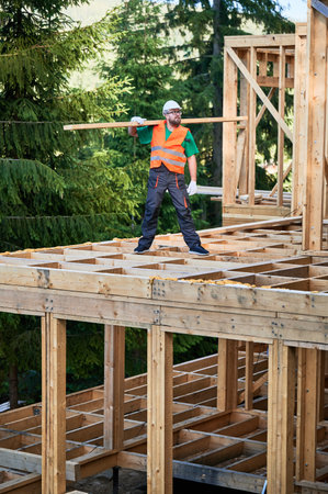 Carpenter constructing wooden-framed house near the woods. Man holds large beam on his shoulder and is dressed in work clothes and helmet. Modern concept of environmentally-conscious building.の写真素材