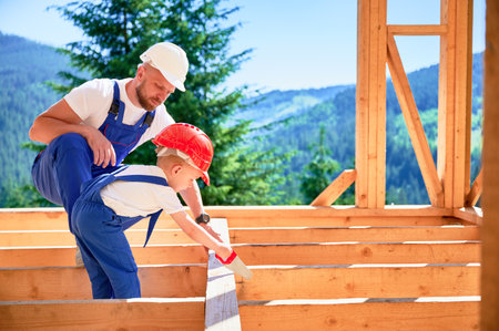 Father with toddler son building wooden frame house. Man teaching the art of manually sawing wood with hand saw on construction site, wearing helmets and blue overalls. Carpentry and family concept.の写真素材