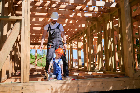 Father with toddler son building wooden frame house. Male worker playing with his son with electric saw on construction site, wearing helmets and overalls on sunny day. Carpentry and family concept.の写真素材