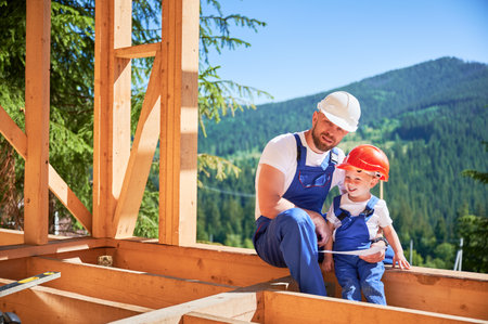 Father with toddler son building wooden frame house in the mountains. Man showing his smiling son the construction plan, wearing helmets and blue overalls on sunny day. Carpentry and family concept.の写真素材