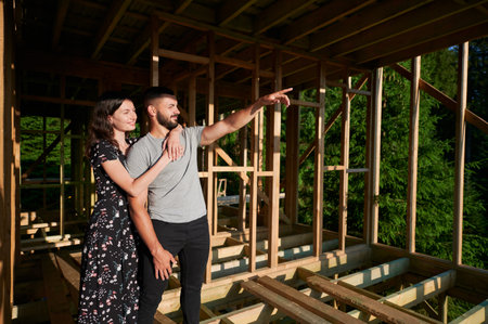 Man and woman inspecting their future wooden frame house nestled in the mountains near forest. Youthful couple at construction site in early morning. Concept of contemporary ecological construction.の写真素材