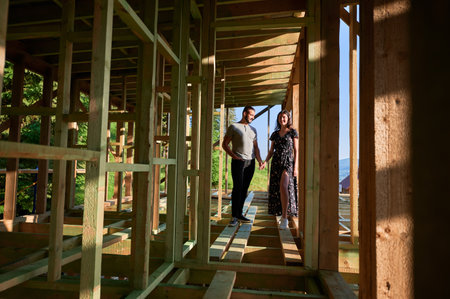 Man and woman inspecting their future wooden frame house nestled near the forest. Youthful couple at construction site in early morning. Concept of contemporary ecological construction.の写真素材