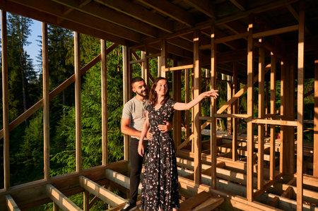 Man and woman inspecting their future wooden frame house nestled near the forest. Youthful couple at construction site in early morning. Concept of contemporary ecological construction.の写真素材